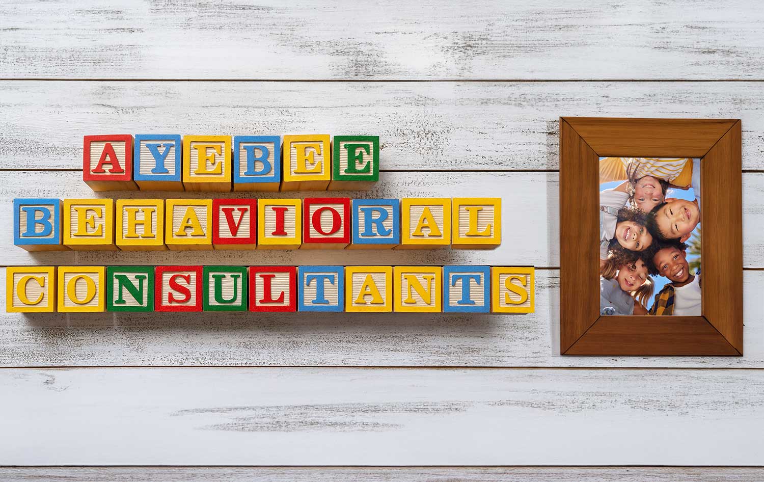 Colorful children's blocks arranged to spell 'Ayebee Behavioral Consultants' next to a framed photo of smiling children, all set against a white wooden background with a bright yellow border.