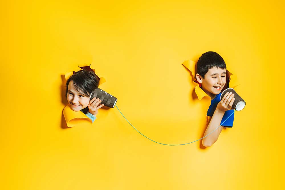 Two smiling children communicating through a tin can telephone against a bright yellow background, symbolizing connection and outreach.