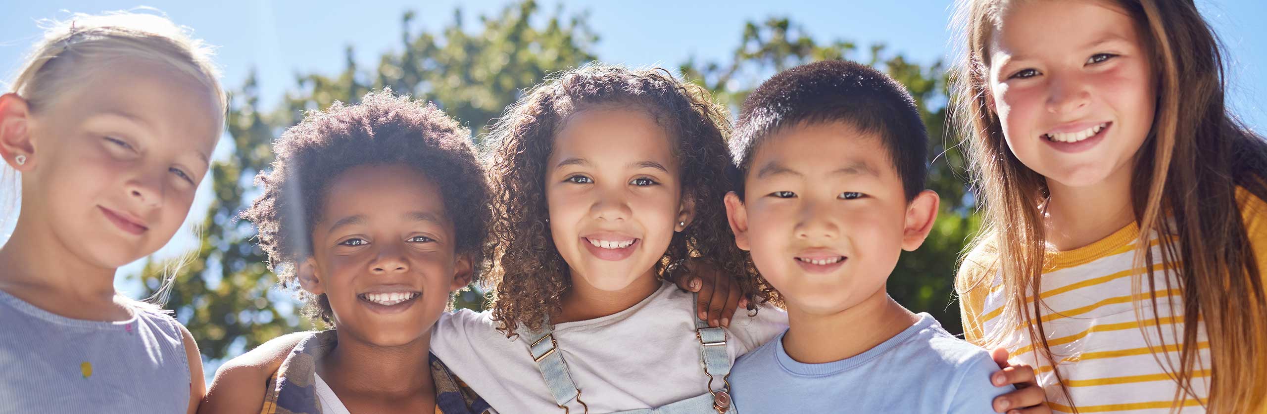 A joyful group of diverse children standing outdoors with their hands raised high, smiling brightly in a sunlit park setting, celebrating friendship and inclusion.